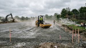 Roller compacting the stone base of a new soccer field under construction with goal frames stacked nearby