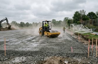 Roller compacting the stone base of a new soccer field under construction with goal frames stacked nearby
